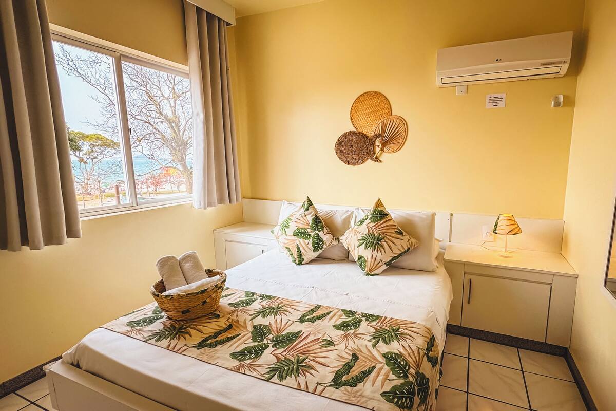 A hotel room with a bed, a bedside table, a lamp, a basket with towels, and a window with curtains.