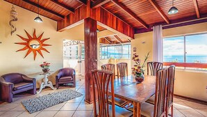 A dining area with a wooden table and chairs, a sun decoration on the wall, and a view of the ocean. - Pousada Vó Regina (Florianópolis)