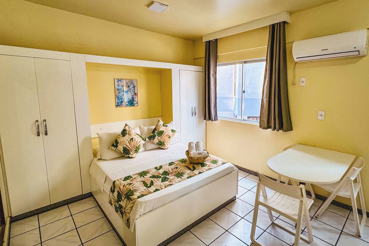 A room with a bed, a small table with chairs, a window with curtains, and an air conditioning unit.