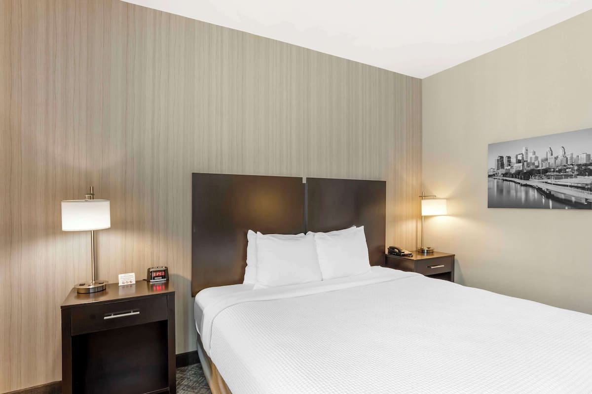 Une chambre d’hôtel avec un grand lit, deux tables de chevet, une petite table, une lampe et un tableau représentant un paysage urbain accroché au mur.
