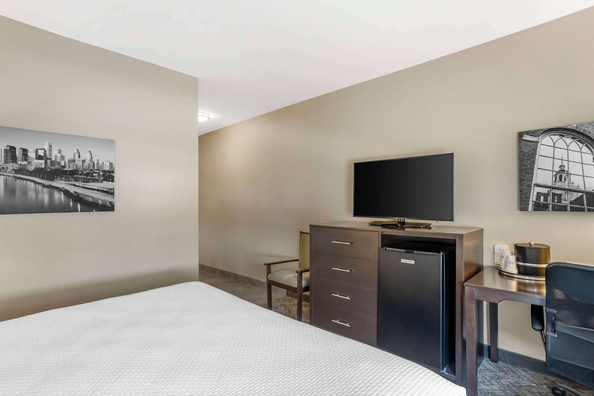 Une chambre d’hôtel comprenant un lit, un bureau avec une chaise, une télévision, un minibar et deux tableaux encadrés accrochés aux murs.
