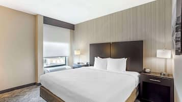 A hotel room with a large bed, a dark headboard, a nightstand with a lamp, and a window with a view of the city.