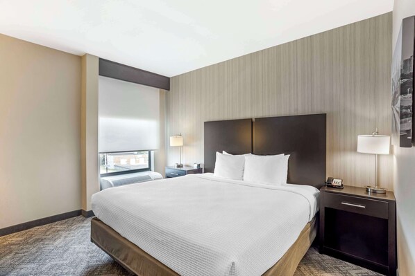 Habitación de hotel con una cama grande, un cabecero oscuro, una mesita de noche con una lámpara y una ventana con vista a la ciudad.