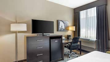 A hotel room with a bed, a desk with a computer, a small refrigerator, a flat-screen TV, a lamp, and a window with curtains.
