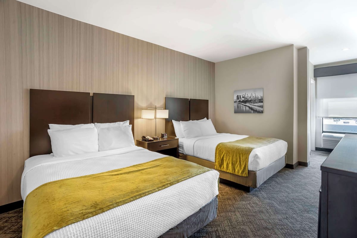 Une chambre d’hôtel avec deux lits, une table de chevet, une lampe, un tableau représentant un paysage urbain accroché au mur et une vue sur l’extérieur.