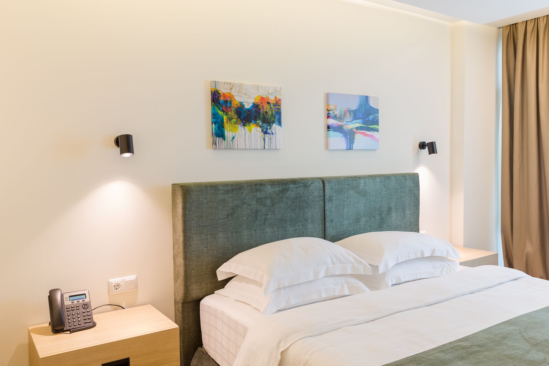 A hotel room with a bed, a bedside table with a phone, and two abstract paintings on the wall.
