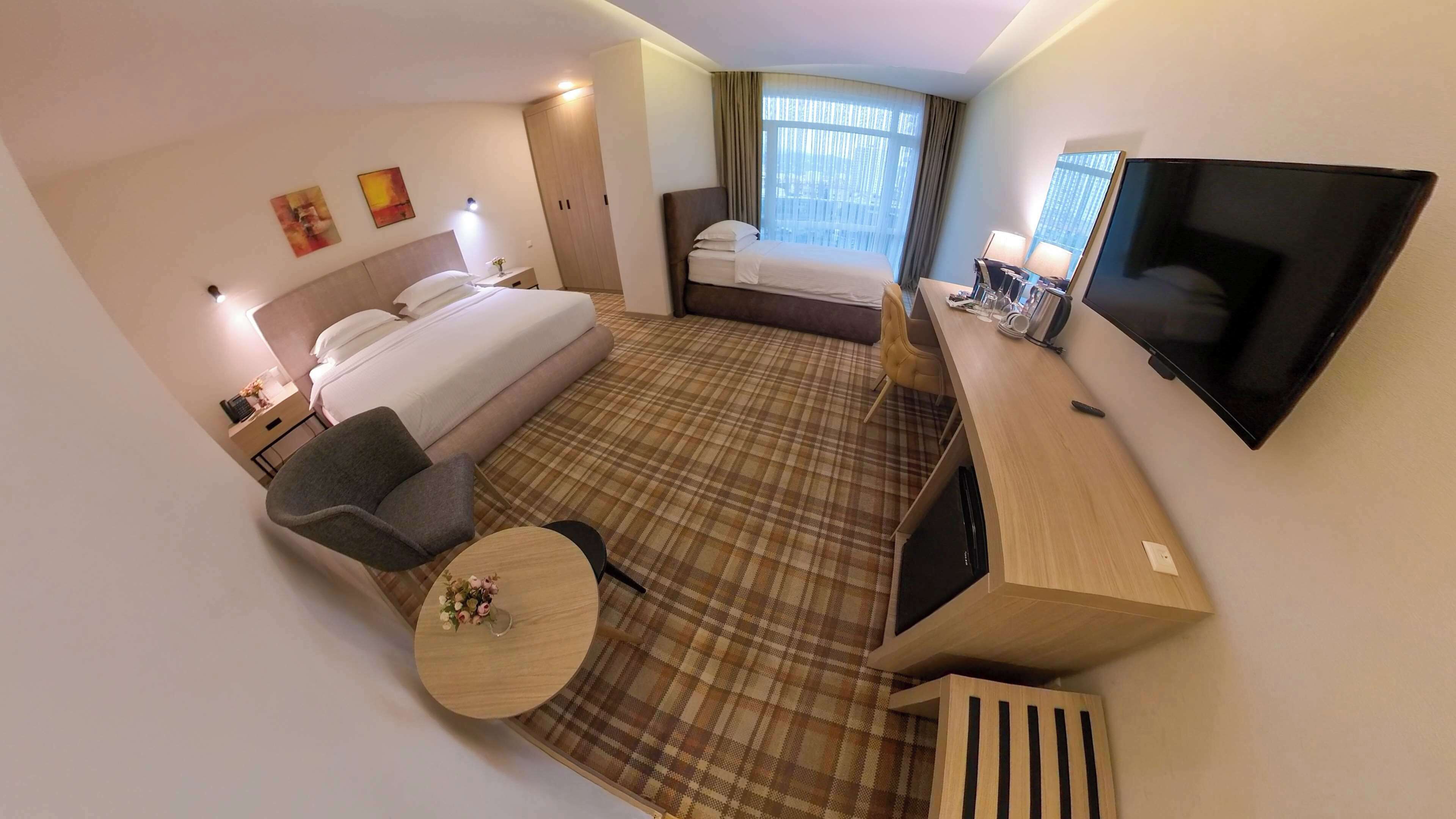Habitación de hotel con cama, escritorio con televisor, silla, mesita con un florero y ventana con cortinas.
