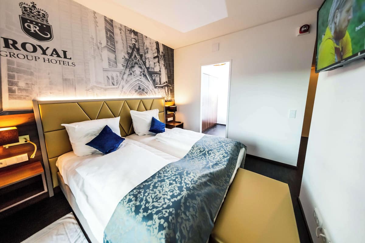 A hotel room with a large bed, a headboard, a wall-mounted TV, and a framed picture of a building.