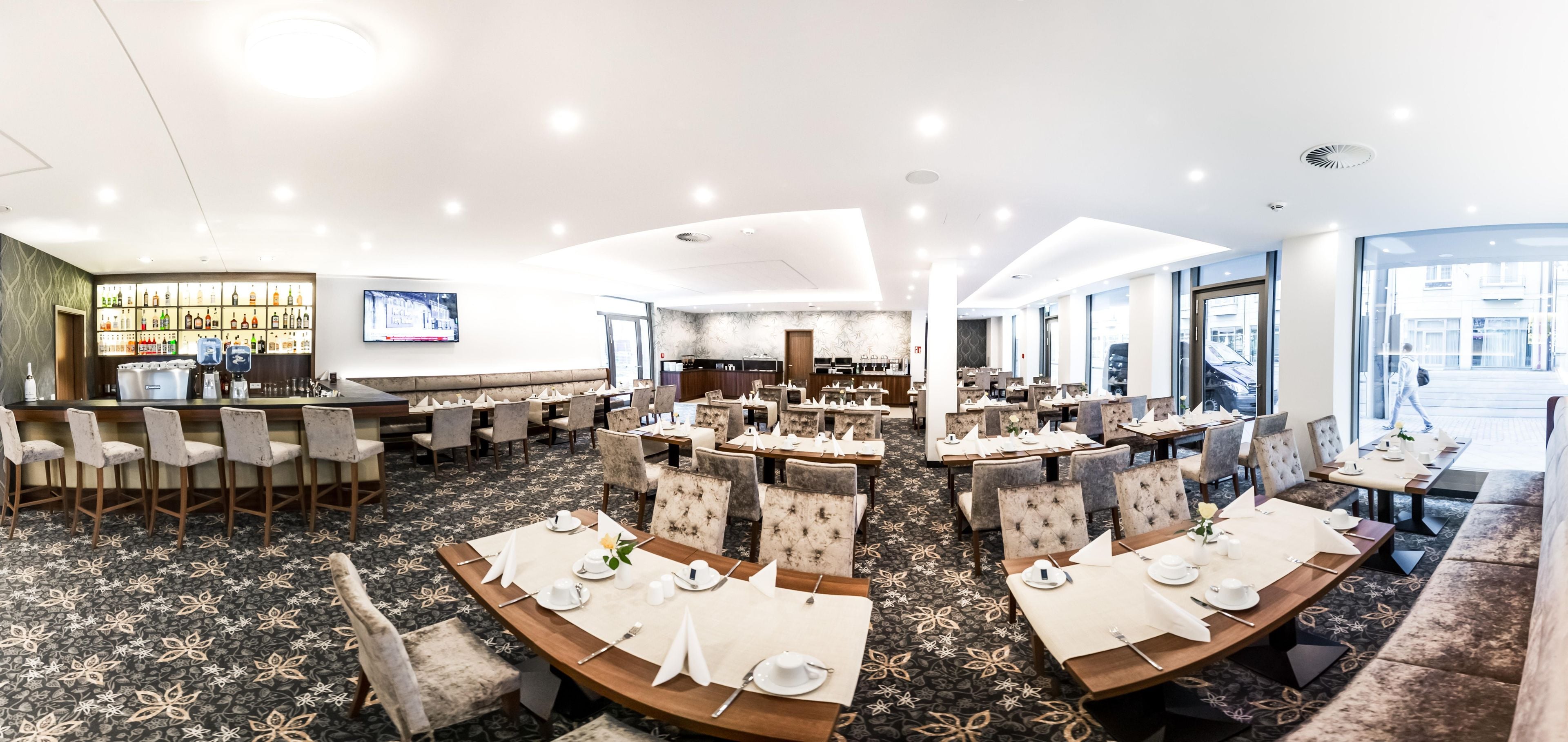 A hotel dining area with multiple tables set for dining, a bar area with a variety of bottles, and a television screen mounted on the wall.