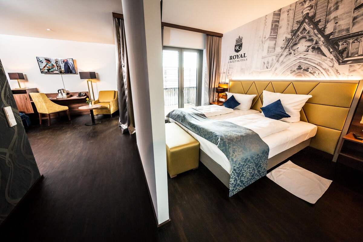 A hotel room with a bed, a desk, a chair, a lamp, and a wall with a cityscape mural.