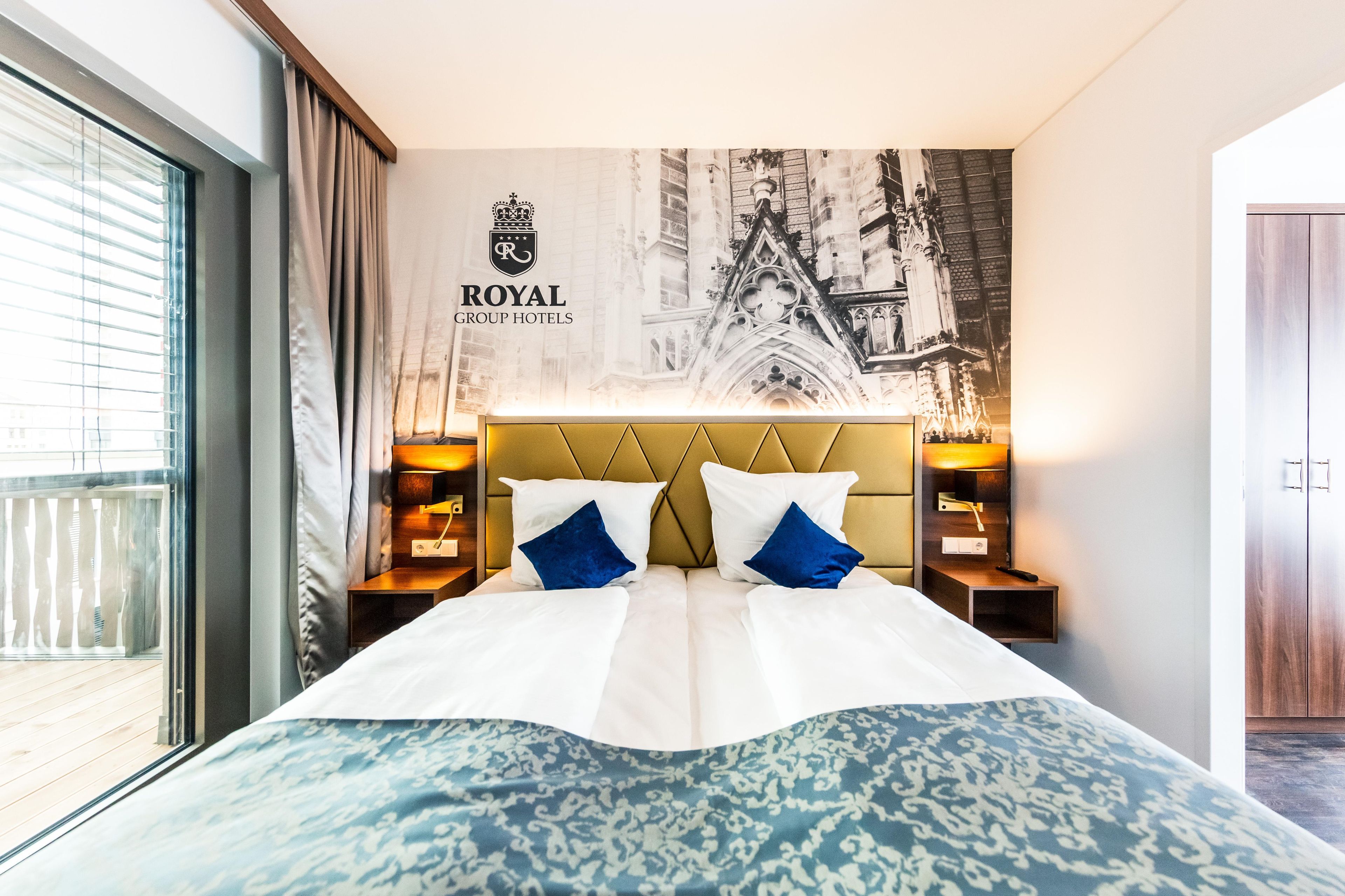 A hotel room with a large bed, a headboard with a crest logo, two bedside tables with lamps, and a large window.