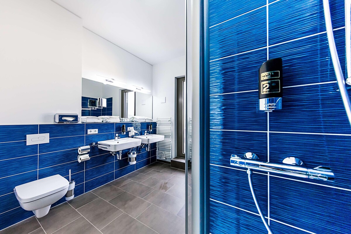A modern bathroom with blue tiles, two sinks, a toilet, and a shower area.