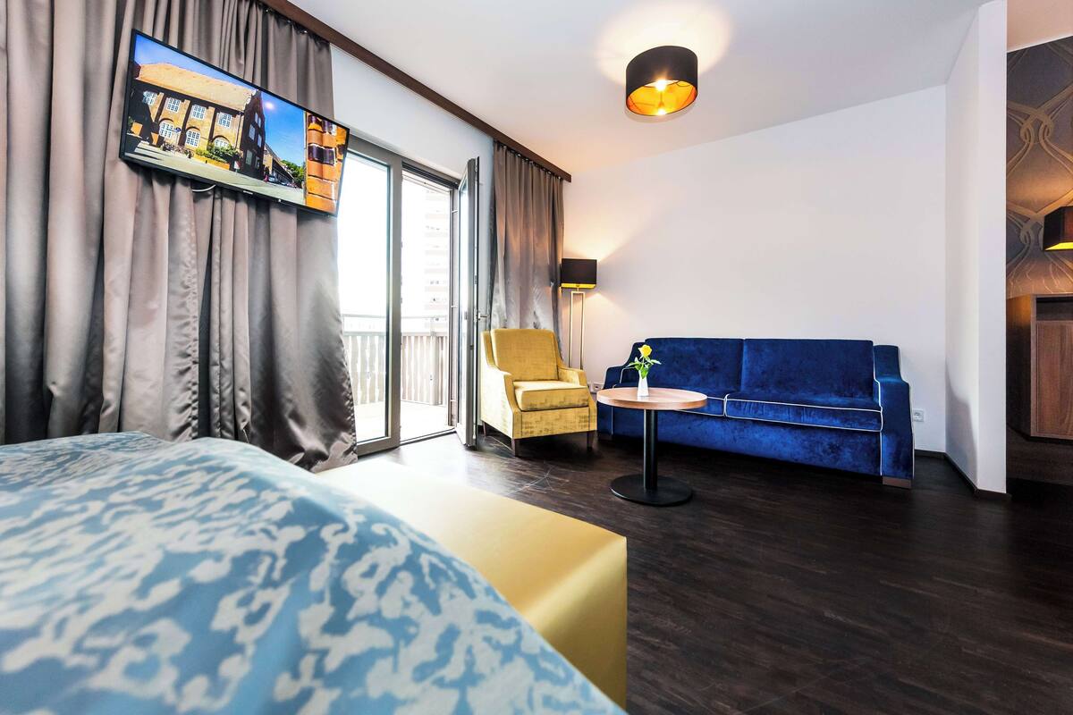 A hotel room with a large bed, a sofa, a chair, a small table, and a TV mounted on the wall.