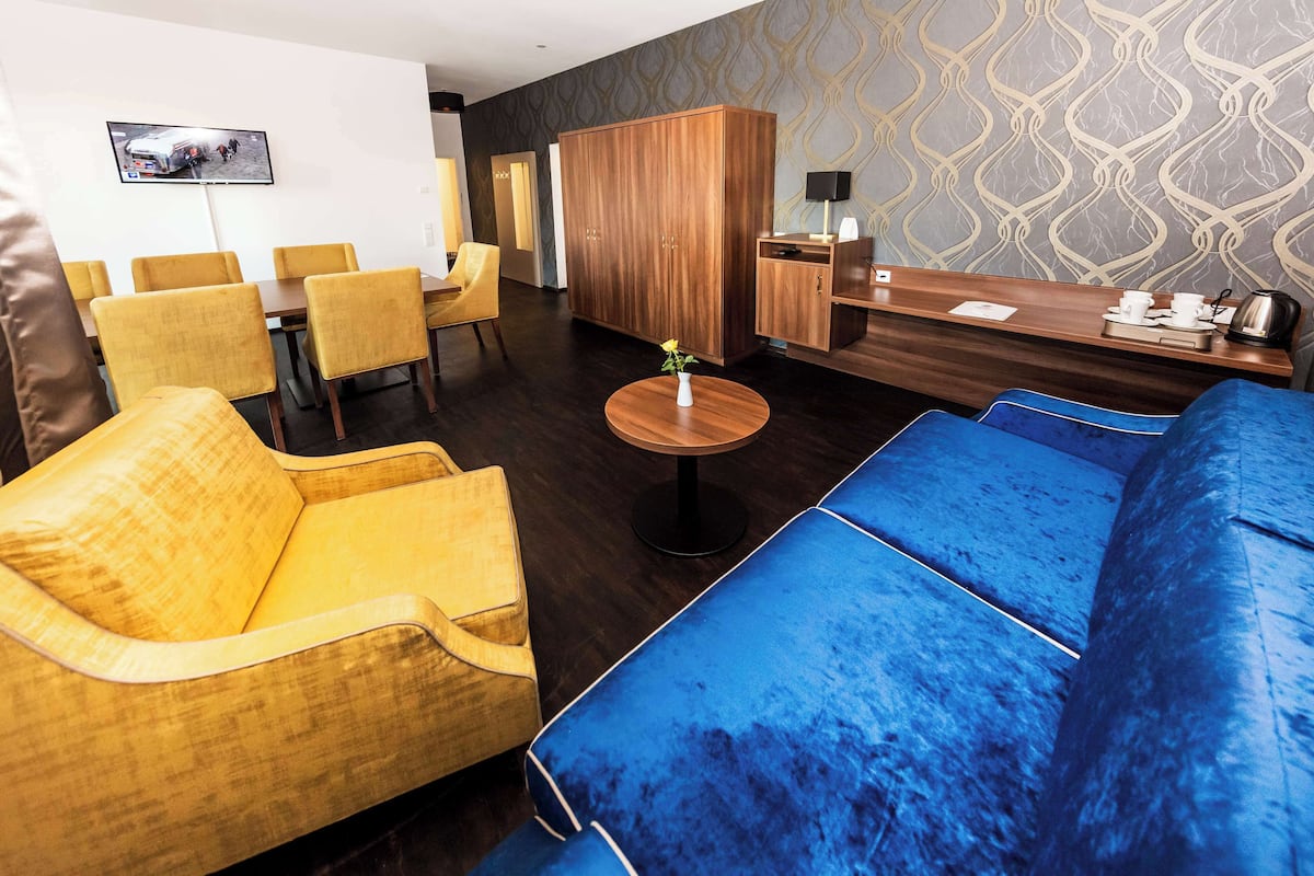 A modern hotel lobby with a blue sofa, yellow armchairs, a wooden reception desk, and a TV mounted on the wall.