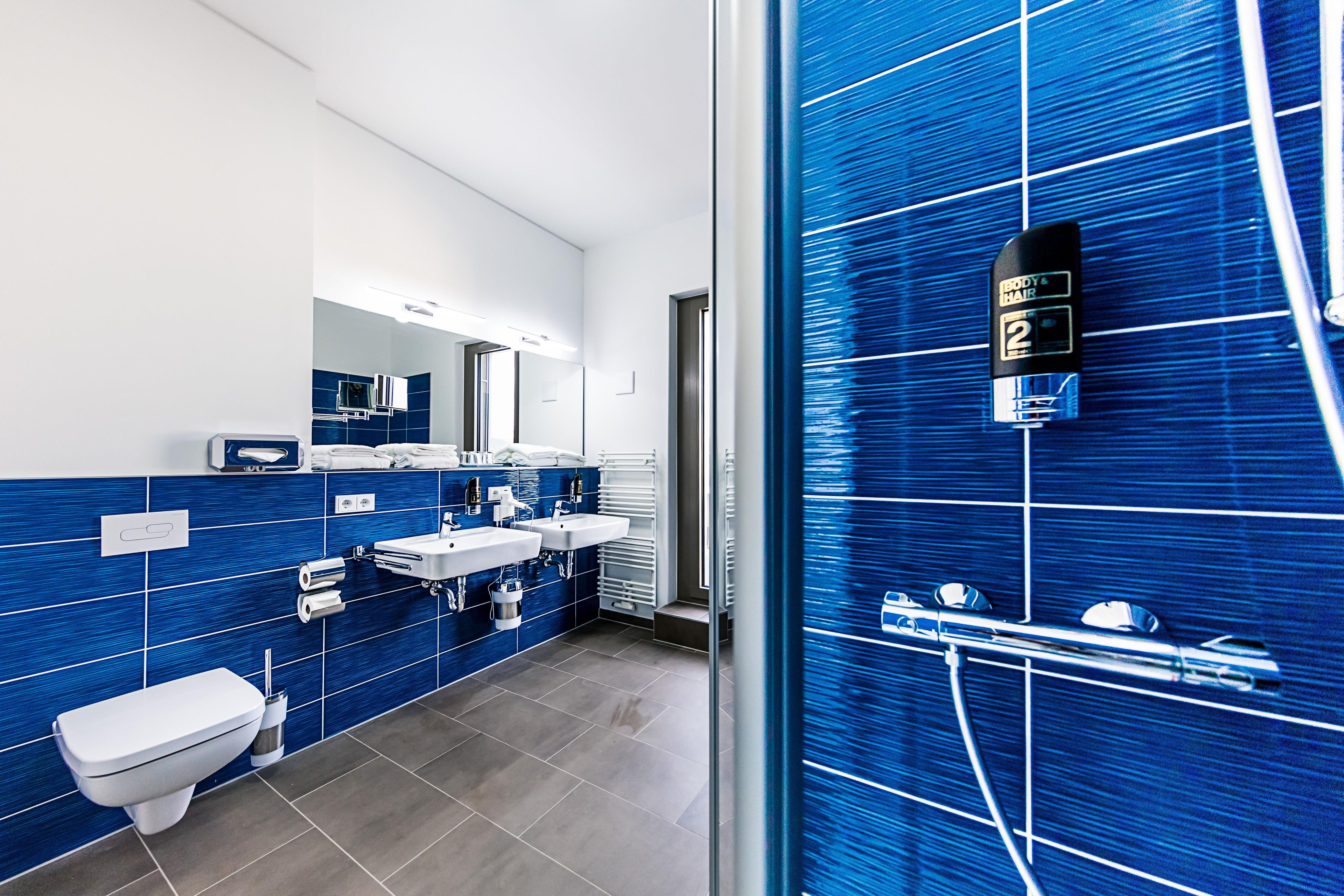 Un baño moderno con azulejos azules, inodoro, dos lavamanos, espejo y zona de ducha.