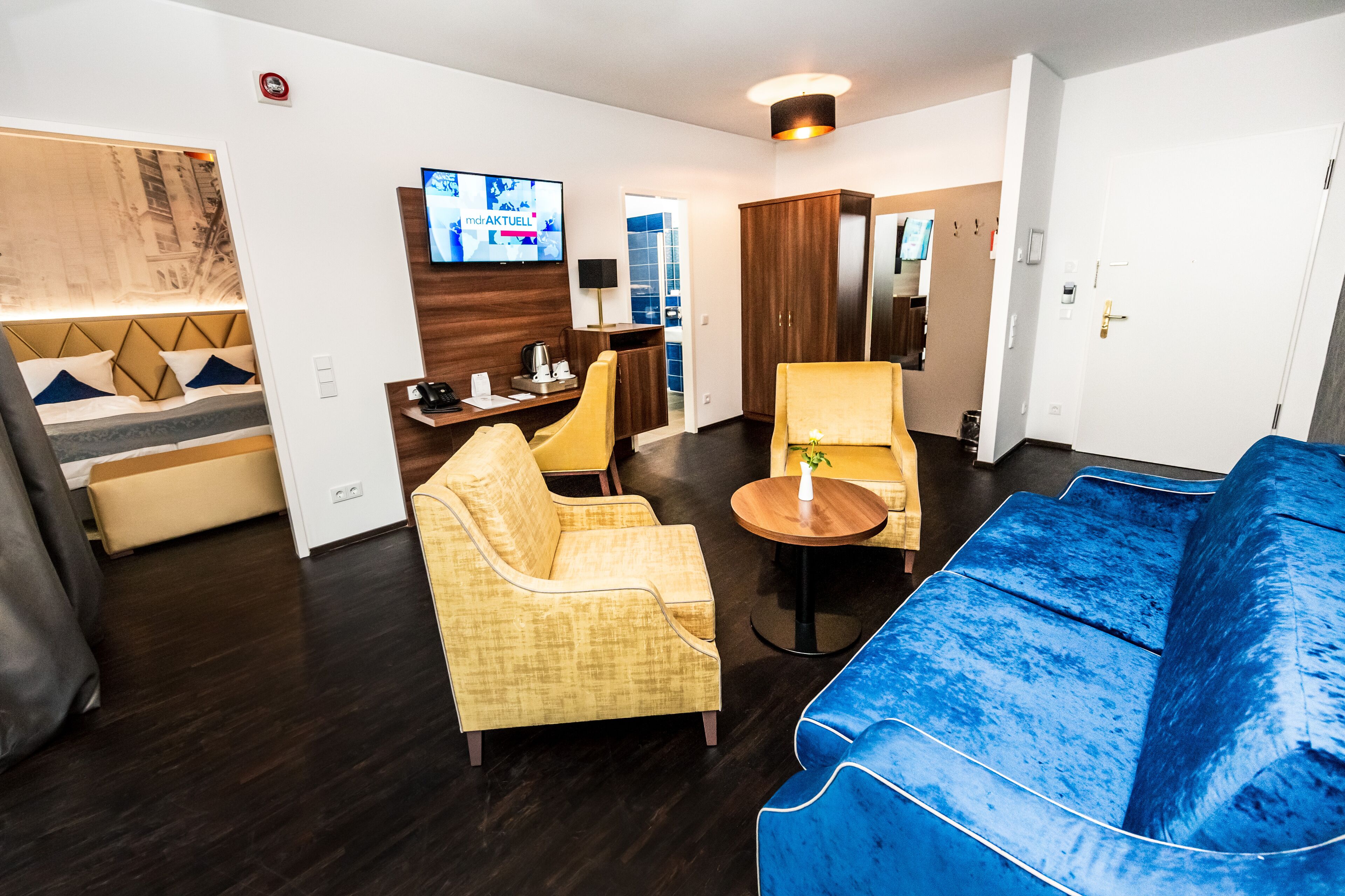 A modern hotel room with a sofa, armchairs, a small table, a TV, and a wardrobe.