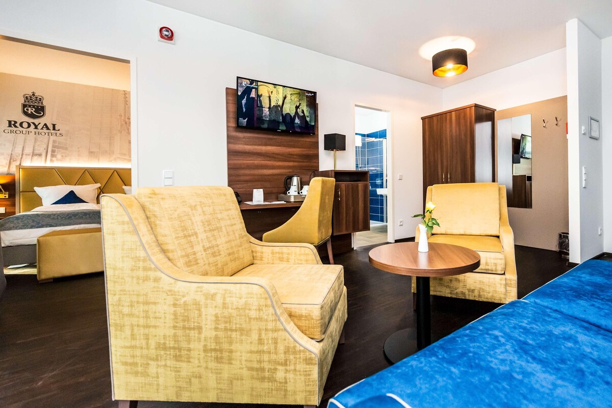 A hotel room with a bed, two armchairs, a sofa, a small table, a TV, and a painting on the wall.