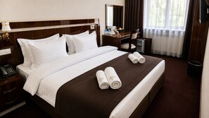 A hotel room with a large bed, a bedside table with a phone, a desk with a chair, and a window with curtains. - Grand Capital Hotel (Tashkent)