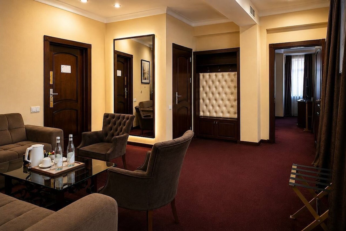 Habitación de hotel con sofá, sillones, una mesita de centro de vidrio, cabecero capitonado y un espejo.