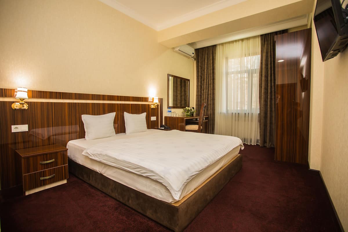 A hotel room with a large bed, wooden headboard, bedside tables, a mirror, a TV, and a window with curtains.