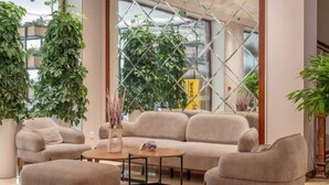 A modern lobby with a chandelier, sofas, a coffee table, and large windows with plants. - Grand Capital Hotel (Tashkent)