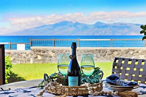 Une table avec une bouteille de vin, deux verres et un panier d’objets décoratifs, le tout placé devant un paysage marin pittoresque.