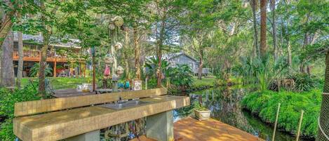 A rustic outdoor dining area with a concrete table and benches, surrounded by lush greenery and a small water feature.