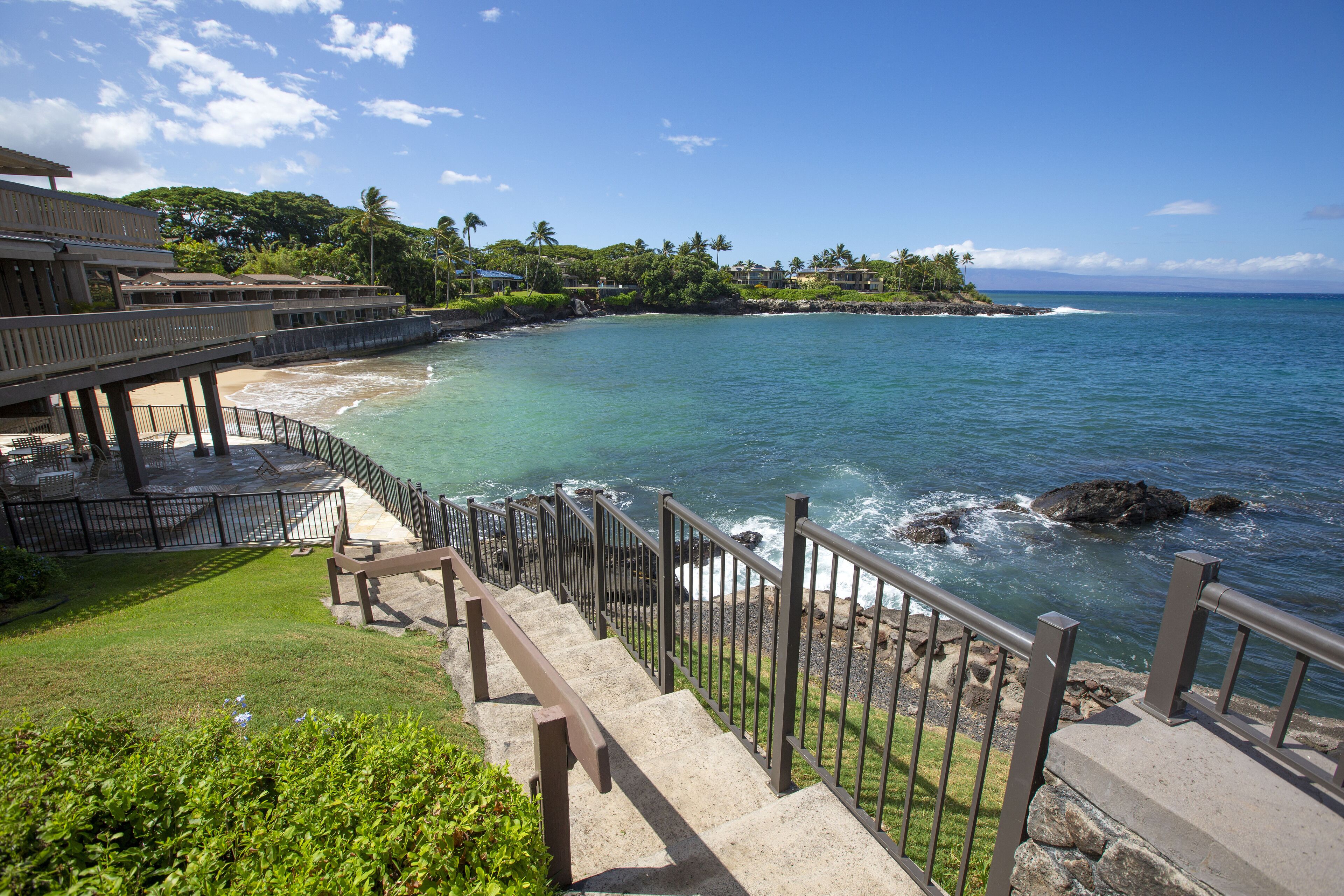A coastal hotel with a wooden deck, a stone staircase leading to a beach, and a view of the ocean.