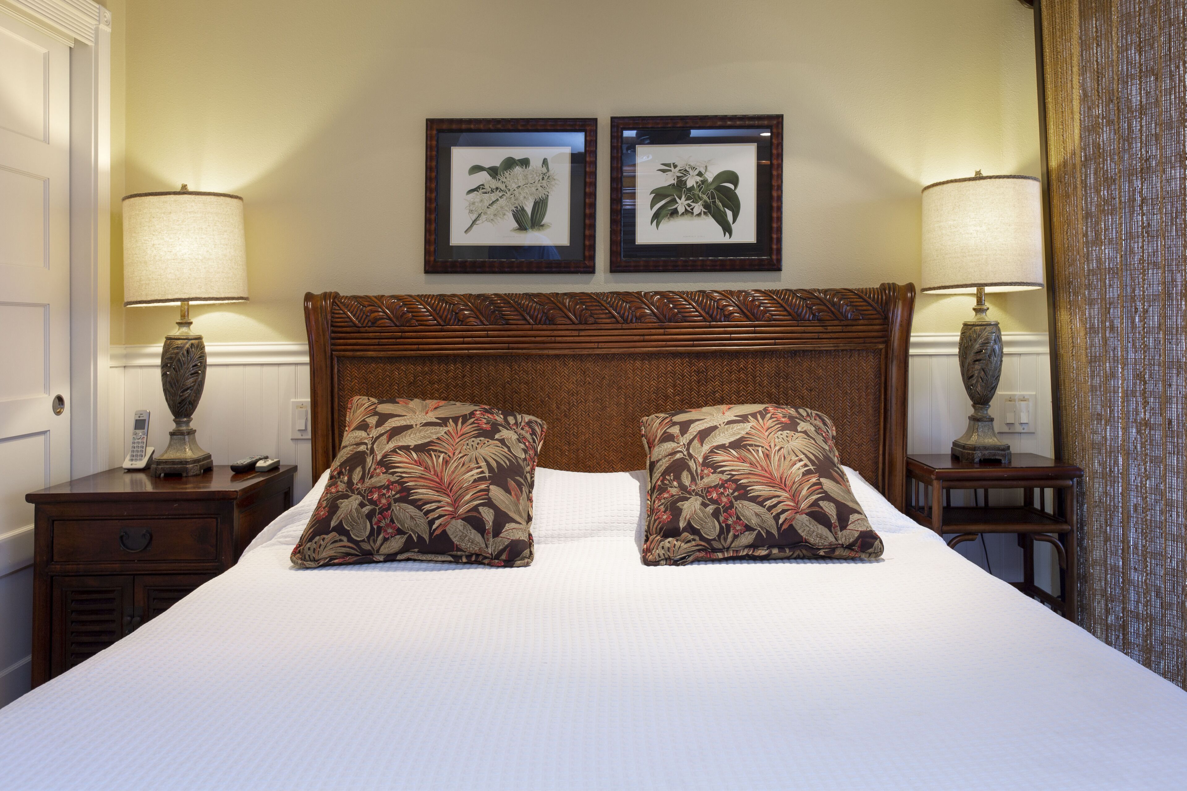 Une chambre d’hôtel avec un grand lit, deux tables de chevet, deux lampes et deux tableaux représentant des motifs floraux, accrochés au mur.
