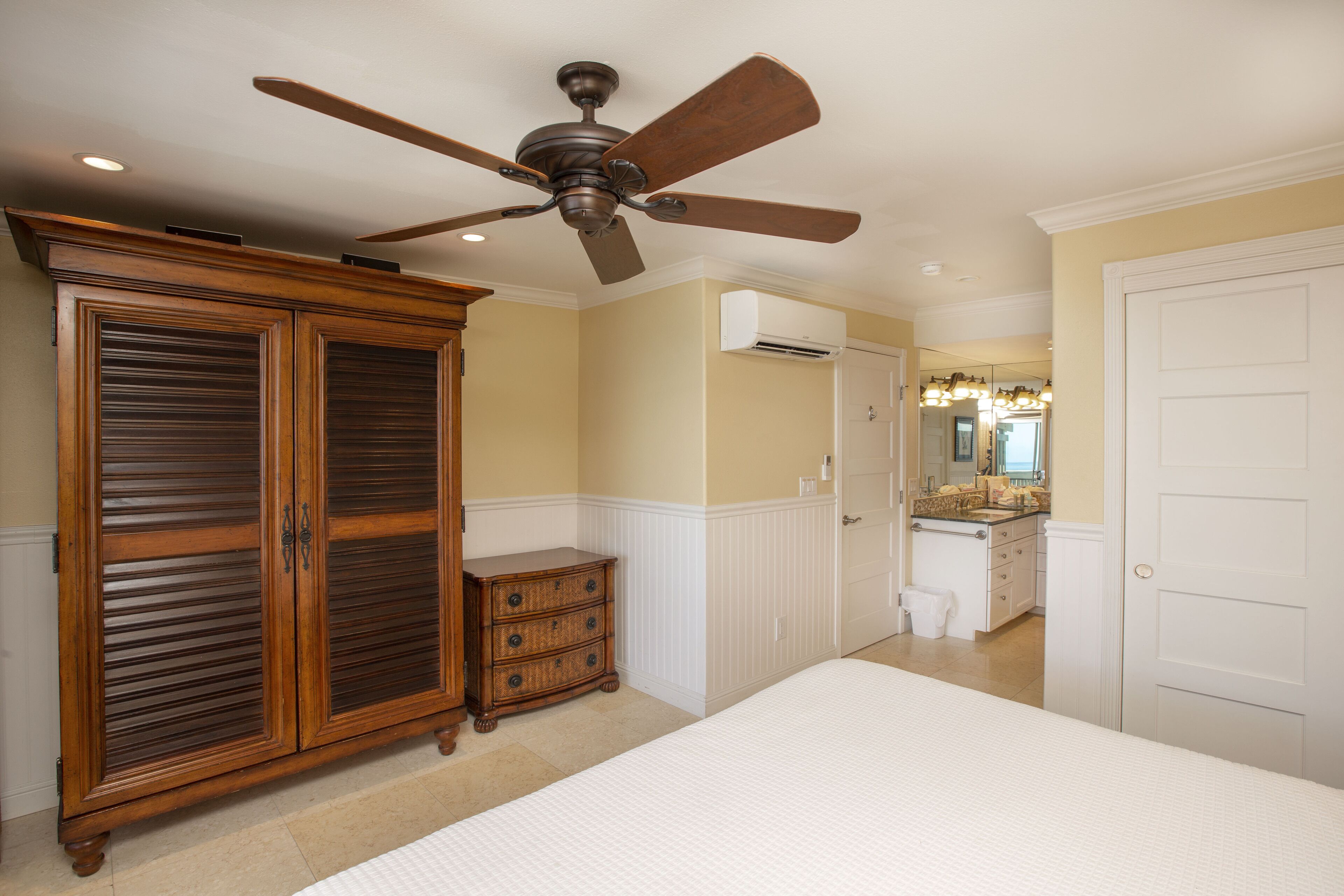 Une chambre à coucher avec un grand lit, une armoire en bois, un ventilateur de plafond et une salle de bain équipée d’un meuble-lavabo et d’un miroir.