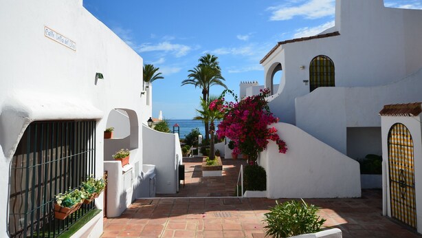 A narrow street with white buildings, a sign reading "Colle Delia dell' Uva," and a view of the sea. - Apartment close to the beach in La Duquesa (Manilva)