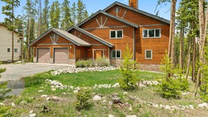 A two-story wooden house with a garage, surrounded by a natural landscape with trees and rocks.