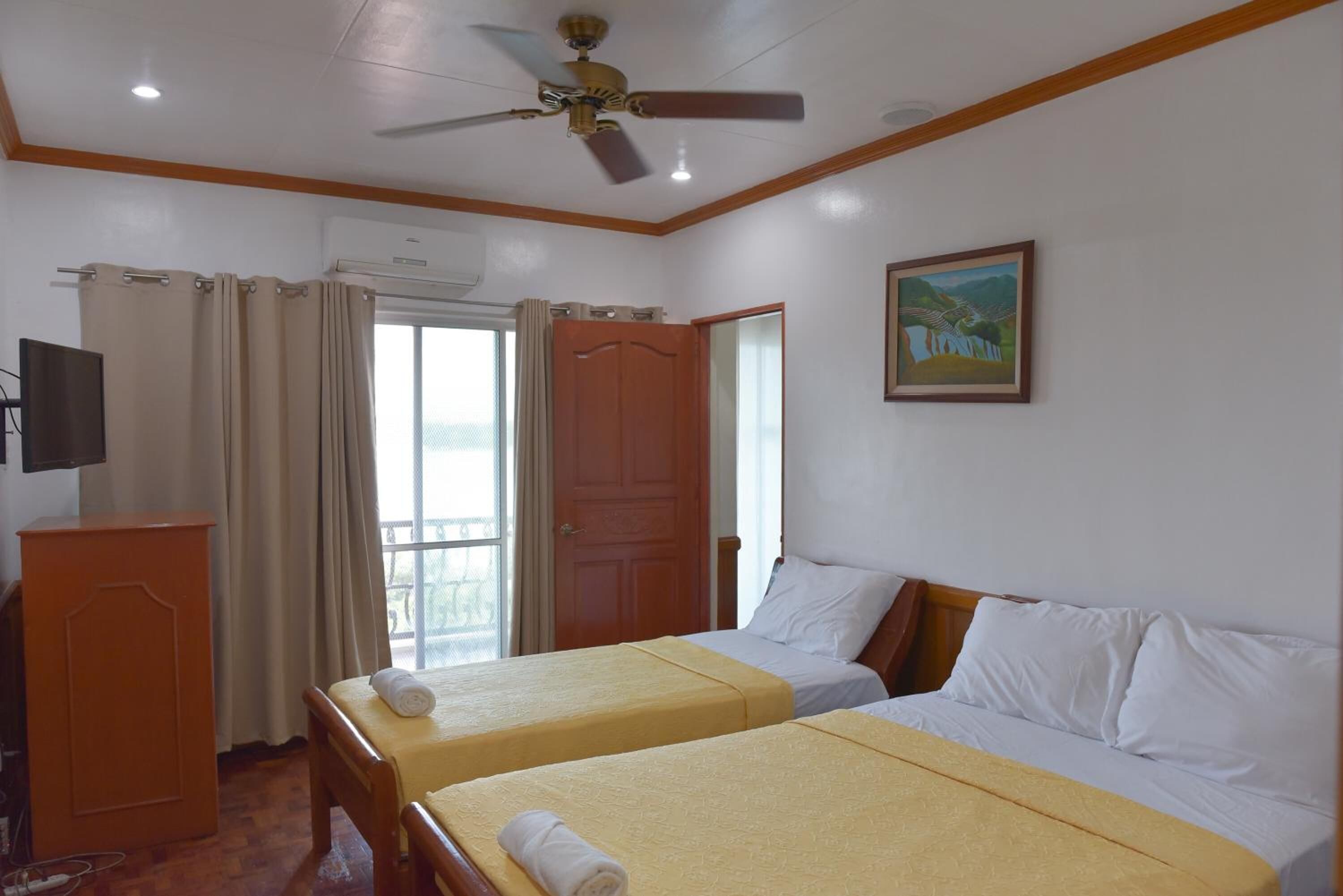 Habitación de hotel con dos camas, un escritorio de madera, ventilador de techo, un cuadro y una ventana con cortinas.