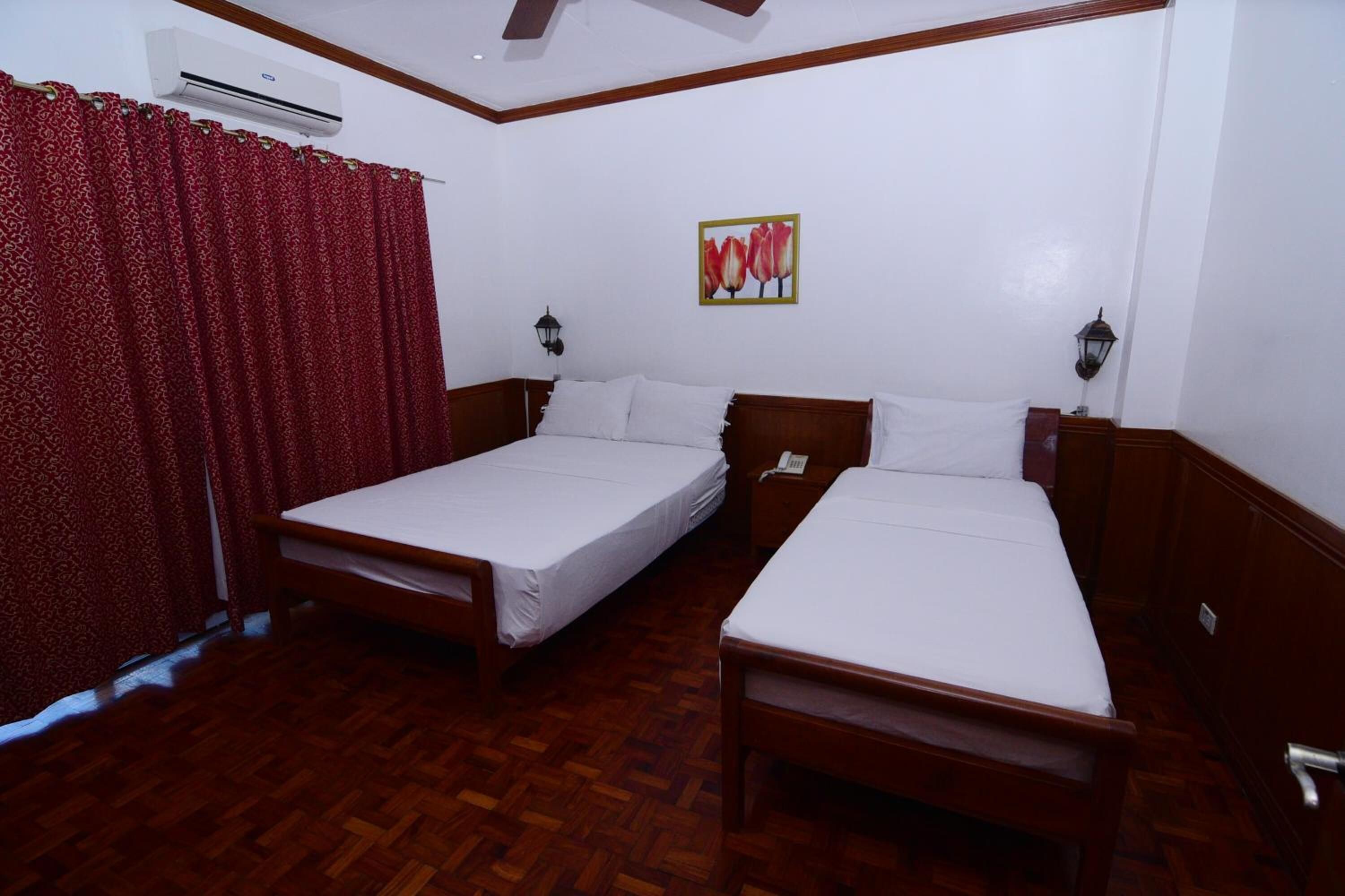 A hotel room with two single beds, wooden flooring, a red curtain, an air conditioning unit, and a wall-mounted lamp.