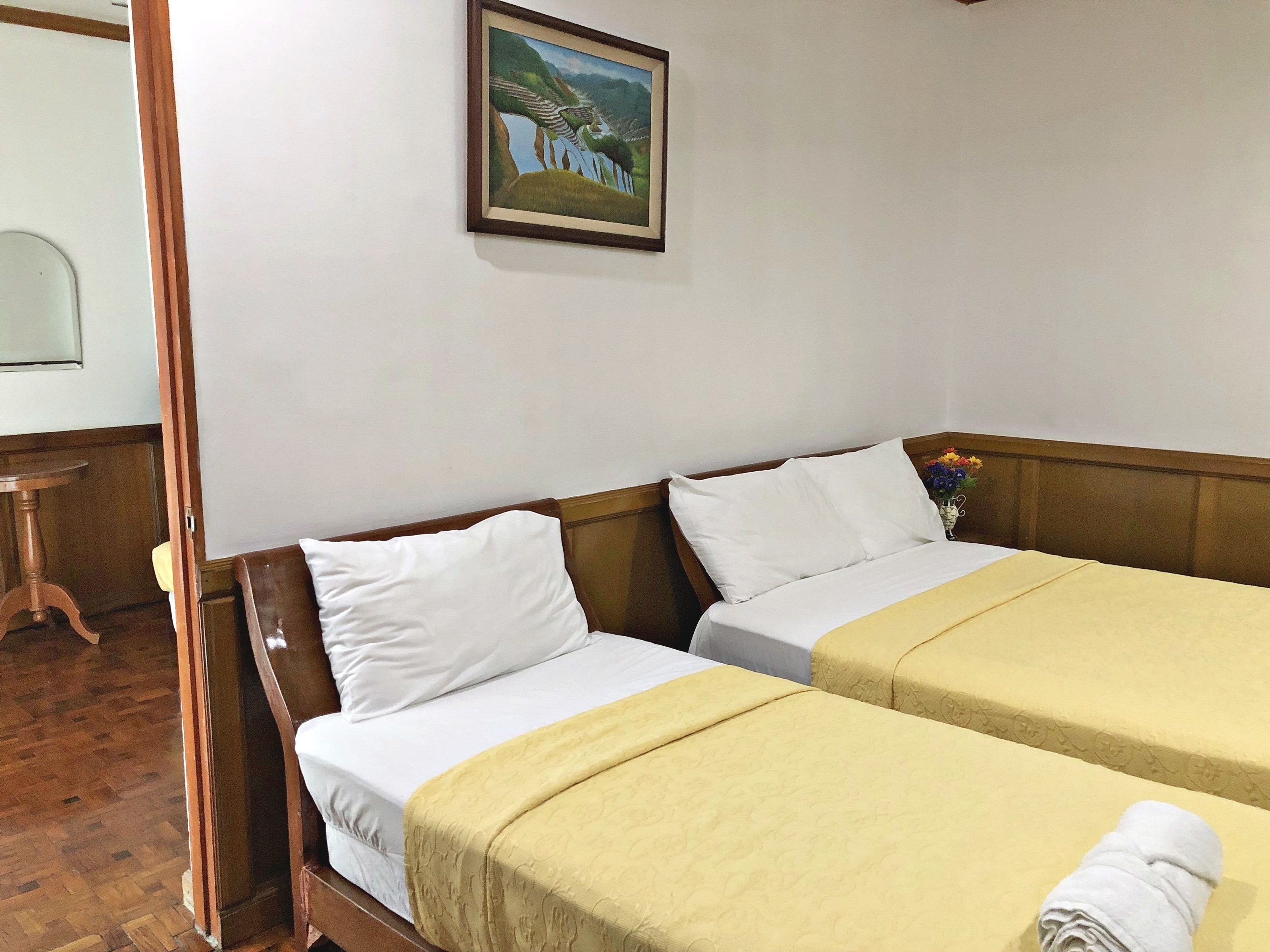 Habitación de hotel con dos camas individuales, cabecero de madera, un cuadro en la pared y una mesita con un florero.