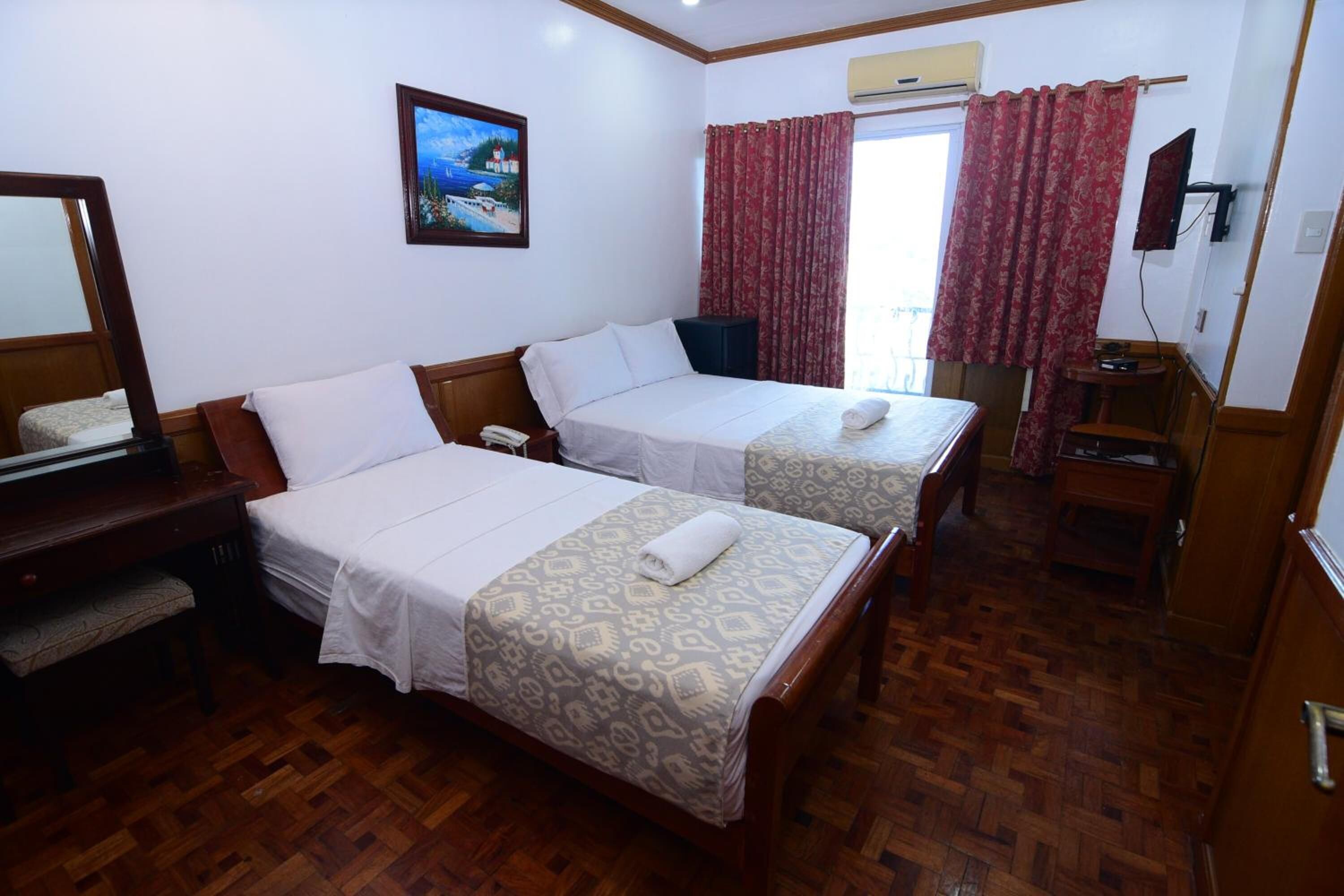 A hotel room with two beds, a desk with a telephone, a mirror, a painting on the wall, and a window with red curtains.