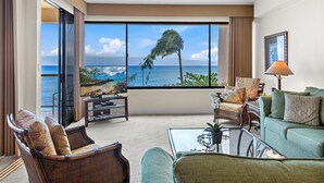 A spacious living room with a large window overlooking a beach and ocean, furnished with a sofa, chairs, and a coffee table.