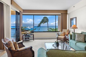 A spacious living room with a large window overlooking a beach and ocean, furnished with a sofa, chairs, and a coffee table.