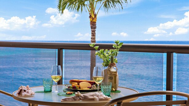 A table with a glass of white wine, a bottle of olive oil, a plate of food, and a small plant on a balcony with a view of the ocean.