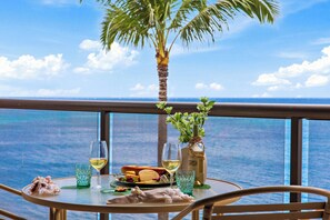 Un tavolo con un bicchiere di vino bianco, una bottiglia d'olio d'oliva, un piatto di cibo e una piccola pianta su un balcone con vista sull'oceano.