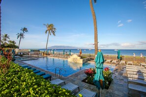 A pool area with lounge chairs and umbrellas, a view of the ocean, and palm trees.