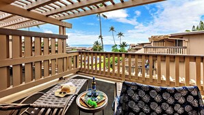 Varanda com grade de madeira, uma mesa com garrafa, copos e um chapéu, vista para uma piscina e palmeiras.