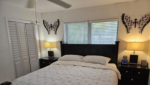 Una cama bien tendida con una colcha estampada, dos mesitas de noche con lámparas, una ventana con persianas y decoración de mariposas en la pared.