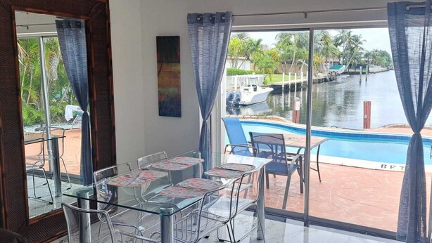 Una habitación con puerta de vidrio que da a una zona de piscina, con una mesa de comedor y sillas, y vistas a un canal con barcos.
