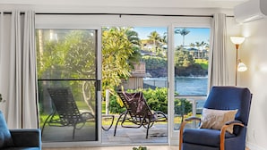 Een kamer met een schuifdeur die uitkomt op een balkon met uitzicht op palmbomen en de oceaan. Er staat een blauwe fauteuil en een rieten stoel op het balkon.