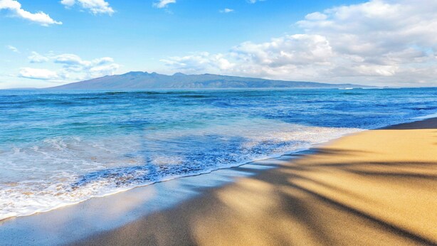 Una playa con aguas azules cristalinas, una orilla arenosa y montañas lejanas bajo un cielo parcialmente nublado.