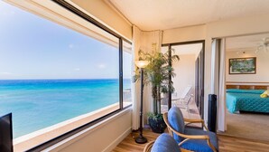 Una habitación con un ventanal grande con vista al océano, una cama con cubrecama verde, una planta en maceta y una silla de mimbre.