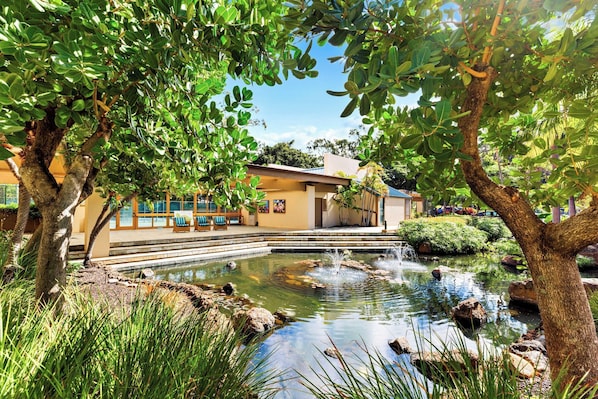 Un jardin paisible avec un étang, un bâtiment doté d’un patio couvert et une végétation luxuriante.