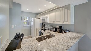 Une cuisine moderne avec des meubles blancs, un plan de travail en granit, un évier, un four à micro-ondes, un grille-pain et une fenêtre avec des stores.