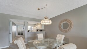 Un coin repas avec une table à plateau en verre et des chaises à revêtement blanc, un luminaire suspendu et une décoration murale en fibres tressées.